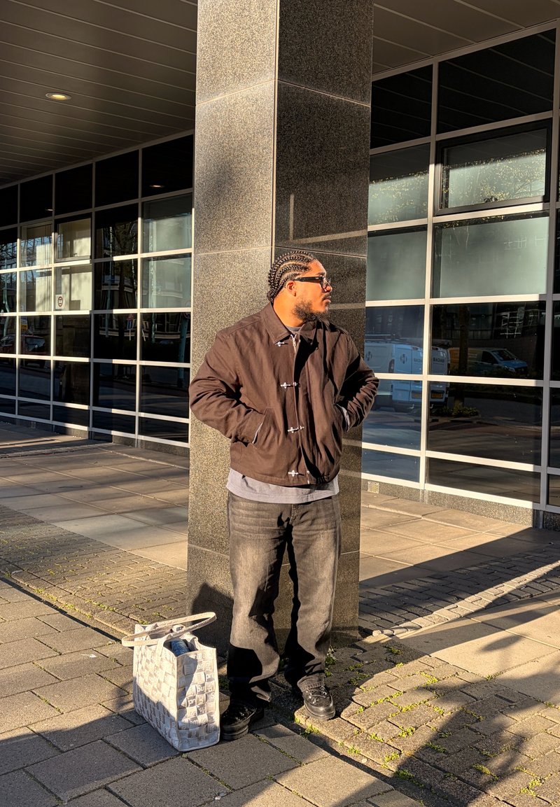 Homme aux cheveux tressés et lunettes de soleil se tient les mains dans les poches près d'un pilier de bâtiment, à côté d'un sac cabas à motifs sur le trottoir.