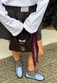 Saco de mão castanho trançado com uma forma curvada, combinado com meias cinzas e mocassins azul claro. Calções pretos às riscas e uma camisa de mangas longas branca.
