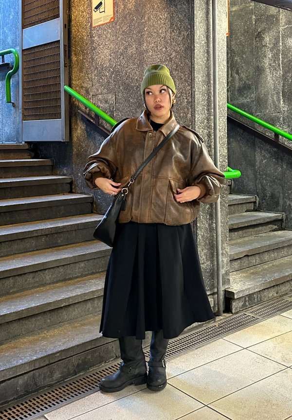 Braune Lederübersize-Jacke, schwarzer Rock, grüner Beanie. Eine schwarze Handtasche und klobige schwarze Stiefel runden das Outfit ab. Treppen im Hintergrund.