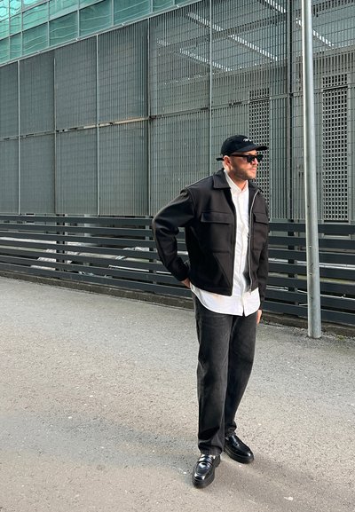 Hombre con gorra negra, gafas de sol, chaqueta negra sobre camisa blanca, pantalones negros y zapatos negros, de pie en una calle adoquinada cerca de una valla de metal.