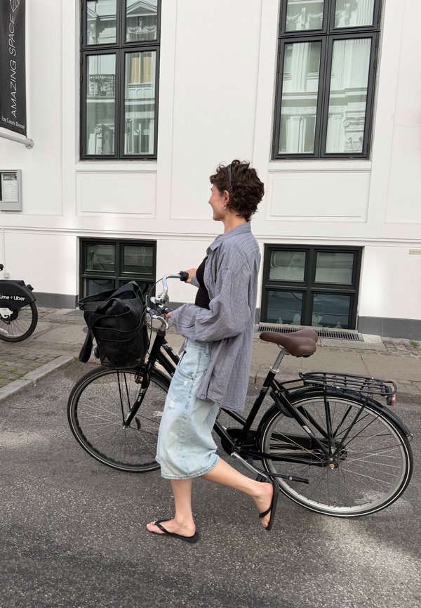 Person, der går med en sort cykel på en byvej ved siden af en hvid bygning med store vinduer.