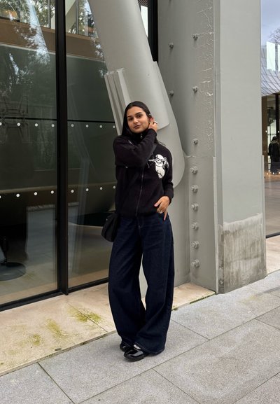 Joven con cabello oscuro que lleva un suéter negro con un gráfico blanco, pantalones anchos oscuros y zapatos negros, de pie junto a un edificio moderno.