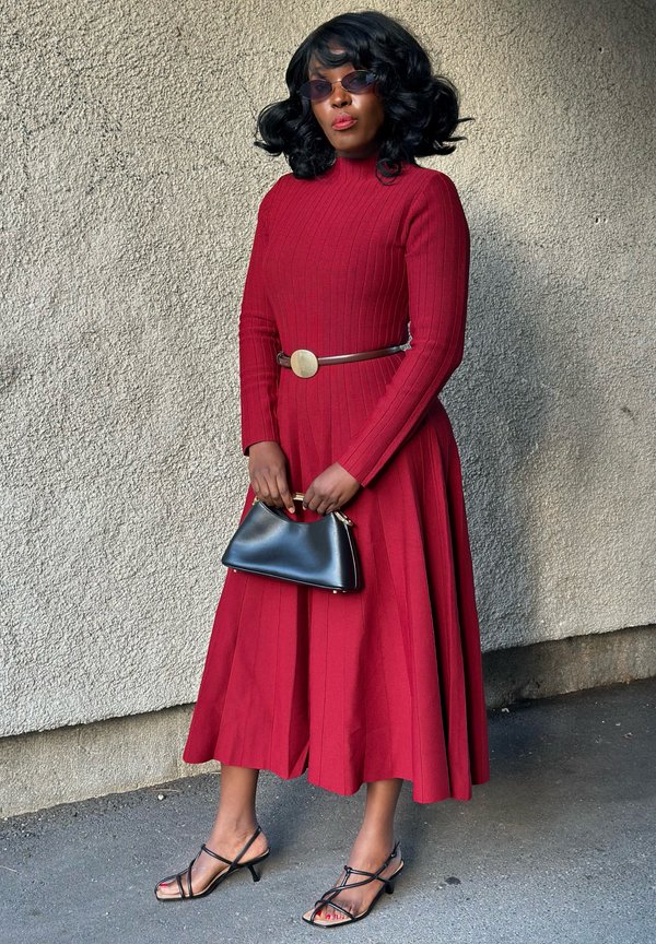 Mujer con un vestido largo rojo acanalado con cinturón, gafas de sol negras, sandalias de tiras y sosteniendo un bolso negro contra una pared texturizada.