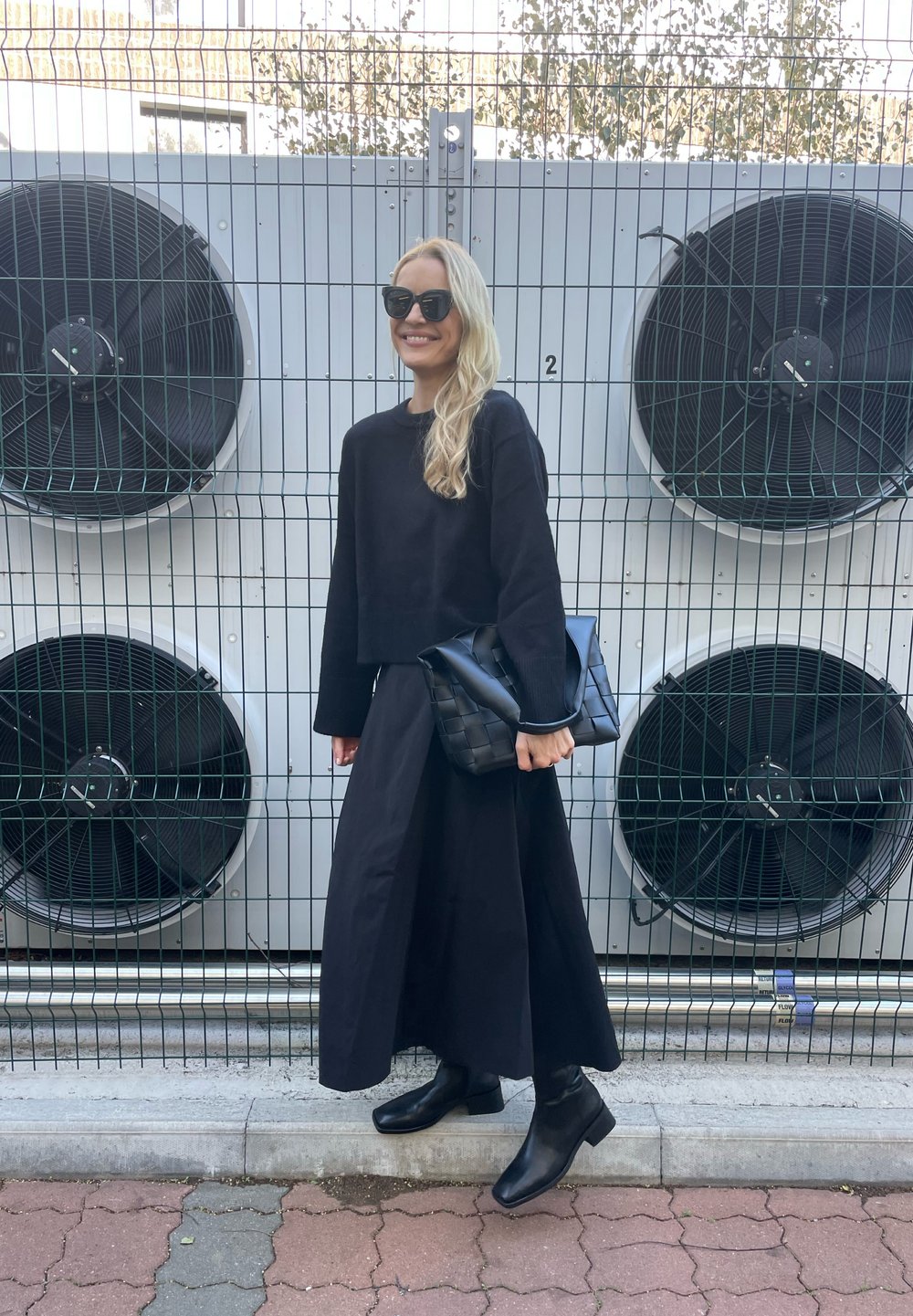 Čierny skrátený sveter, dlhá čierna sukňa a čierne členkové topánky; drží štruktúrovanú čiernu kabelku; veľké priemyselné ventilátory v pozadí.