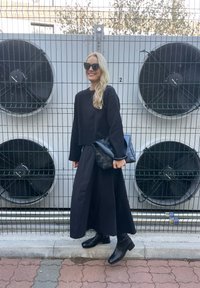 Čierny skrátený sveter, dlhá čierna sukňa a čierne členkové topánky; drží štruktúrovanú čiernu kabelku; veľké priemyselné ventilátory v pozadí.