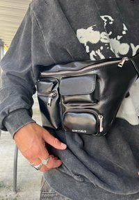 Borchia in finta pelle nera con più tasche con cerniera, forma ovale e hardware in argento. Accentuata con un logo sul davanti.