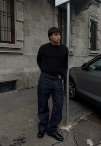 Sort sweater, løse mørke denimbukser og sorte sko. Står op ad en pæl på en struktureret fortov med en stensbygning i baggrunden.