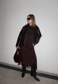 Femme portant une veste en cuir marron foncé, une jupe assortie à ourlet en dentelle, des bottes montantes, des lunettes de soleil noires, et tenant un sac à main texturé rouge foncé.
