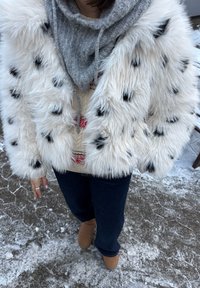 Personne portant une veste en fausse fourrure blanche à taches noires, un sweat à capuche gris, un pantalon bleu marine et des bottes marron, debout sur un sol enneigé.