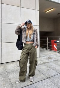 Valehäärtsest jope kapuutsiga, hall lühikeste varrukatega topp, laiad oliivrohelised cargo püksid, hallid spordialused, ja must müts. Must kott on õlal.