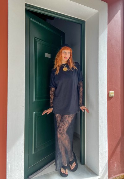 Persona con cabello rojo que lleva una camisa negra de gran tamaño y una falda transparente de encaje, de pie en una puerta con una puerta verde y paredes rojas y blancas.