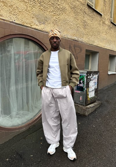 Hombre con chaqueta beige, camisa blanca, pantalones claros y holgados, zapatillas blancas y pañuelo estampado en la cabeza, de pie en una calle urbana junto a una ventana redondeada.