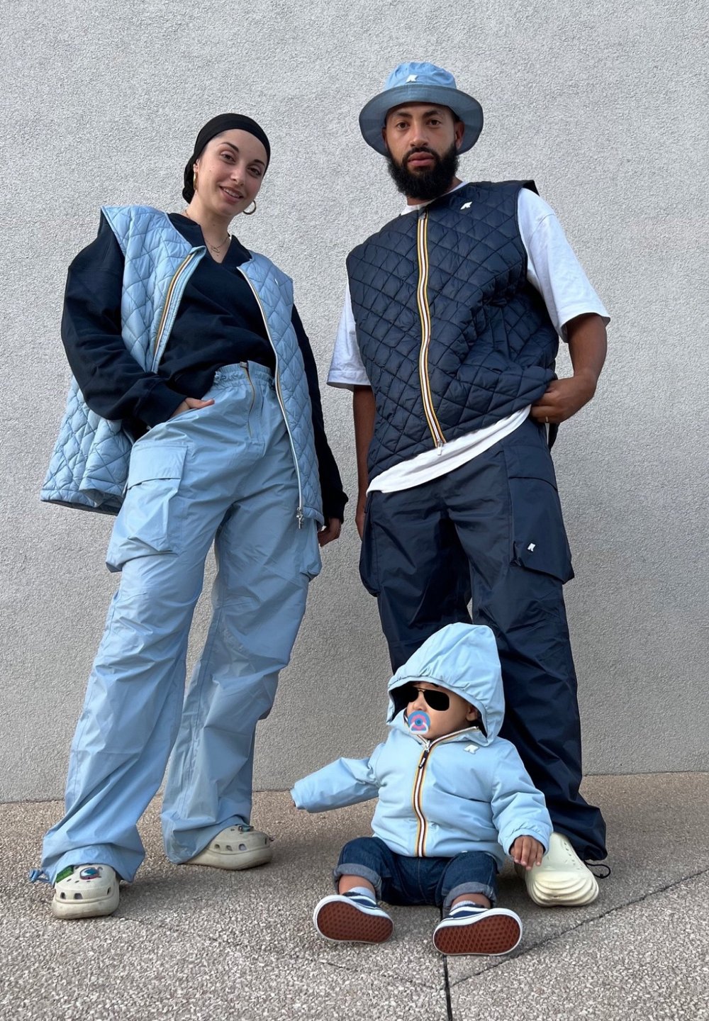 Two individuals are wearing matching blue quilted vests and cargo trousers. A doll dressed in a blue jacket and trousers sits on the ground, wearing trainers.