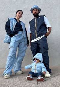 Two individuals are wearing matching blue quilted vests and cargo trousers. A doll dressed in a blue jacket and trousers sits on the ground, wearing trainers.