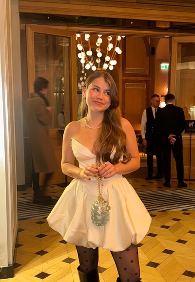 Mujer con vestido blanco sin tirantes y medias de lunares, sosteniendo un bolso adornado con perlas, de pie en una sala iluminada con luz cálida y con personas al fondo.