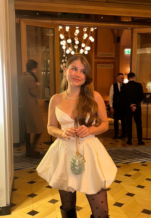 Femme en robe blanche sans bretelles avec des collants à pois tenant un sac orné de perles, debout dans une pièce éclairée chaleureusement avec des personnes en arrière-plan.