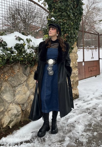 Mujer en un entorno nevado al aire libre que lleva un abrigo negro con ribete de piel sintética, vestido vaquero con un cinturón ancho decorativo, botas negras, boina y un bolso negro.