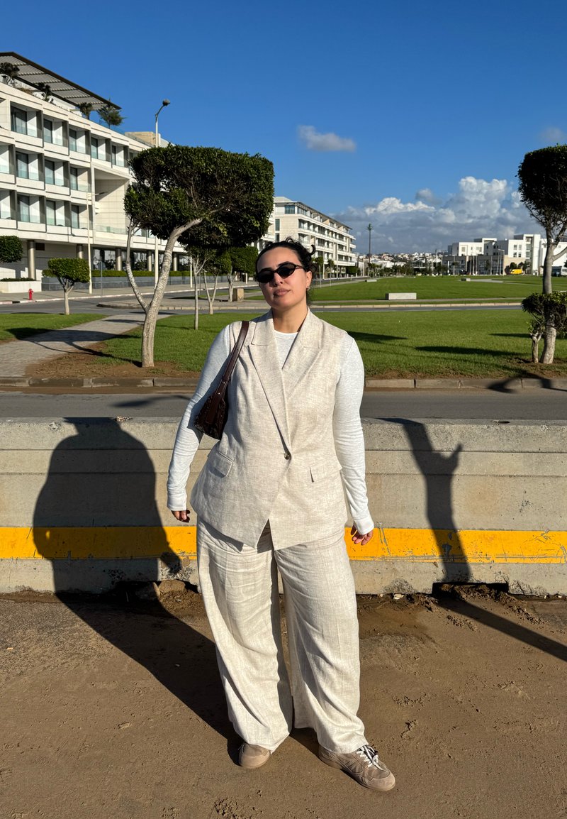 Persona che indossa un gilet beige e pantaloni ampi, camicia bianca a maniche lunghe, occhiali da sole, in piedi su una strada urbana soleggiata con alberi potati e edifici moderni.