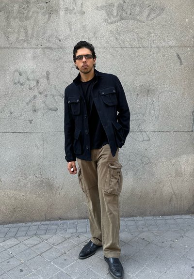 Hombre joven con chaqueta negra, camisa negra, pantalones cargo beige, gafas de sol y zapatos negros, de pie junto a una pared de hormigón gris con grafitis.