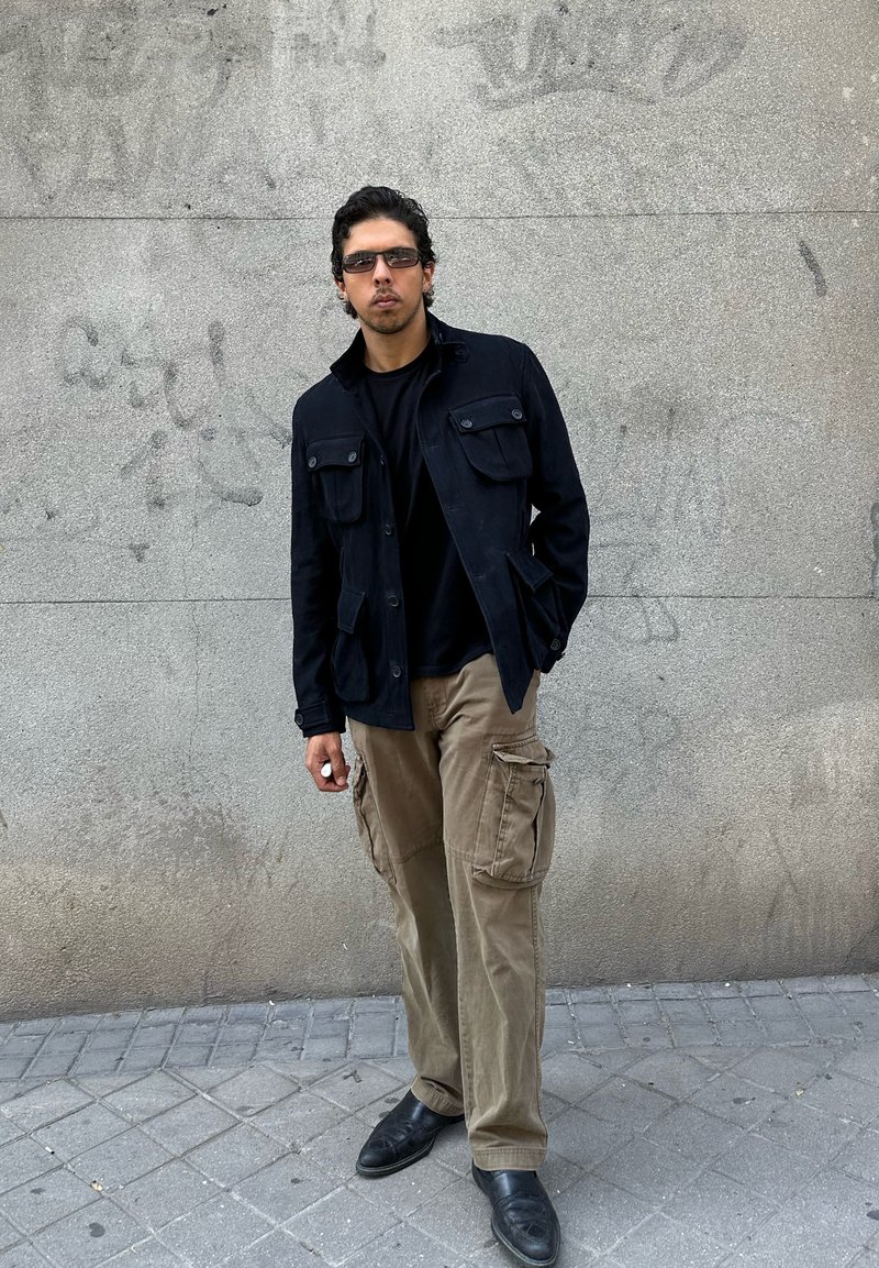 Junger Mann mit schwarzer Jacke, schwarzem Hemd, beigen Cargohosen, Sonnenbrille und schwarzen Schuhen steht vor einer grauen Betonwand mit Graffiti.