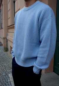 Suéter de malha azul claro com decote redondo canelado, tecido texturizado e punhos canelados; combinado com calças escuras e um fundo neutro.