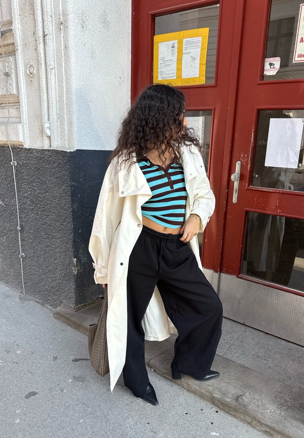 Kvinde med krøllet hår i striped crop top, hvid lang jakke, sorte brede bukser og spidse sko står på fortovet ved en rød dør.