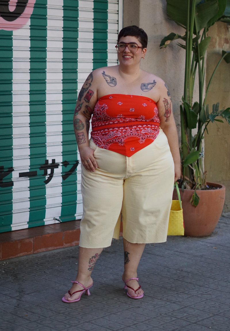 Personne souriante aux cheveux courts et lunettes porte un haut bandana rouge, un short beige, des sandales roses et tient un sac jaune, debout près de plantes en pot.