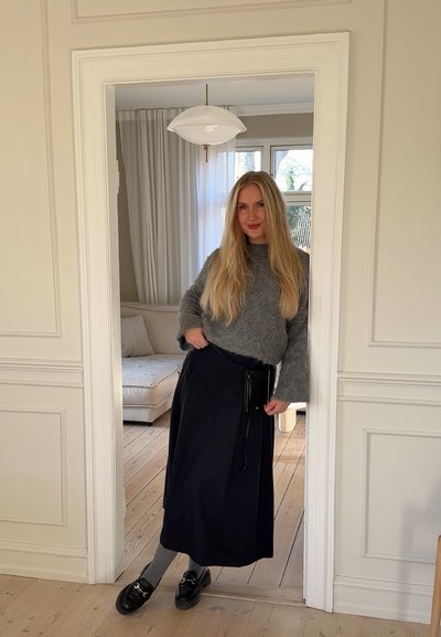 Suéter gris de felpa, falda midi negra, medias gris y zapatos negros. Ambiente interior con suelo de madera clara y una lámpara colgante simple.