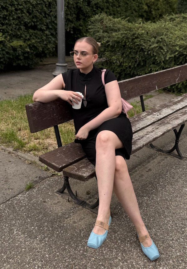 Femme portant une robe noire et des chaussures bleues assise sur un banc en bois dans un parc, tenant une tasse blanche, avec de la verdure et un lampadaire à proximité.