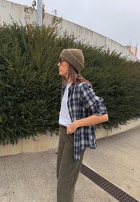 Cămașă din flanel în carouri, în nuanțe de bleumarin și alb, purtată deschisă peste un tricou alb, asortată cu pantaloni din catifea în culoarea verde măsliniu și o bonetă maro.