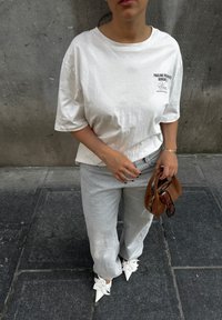Fehér, oversized pólót, világosszürke farmert és masnis fehér, hegyes orrú cipőt viselő nő áll a burkolt úton, kezében egy barna kézitáskával és napszemüvegével.