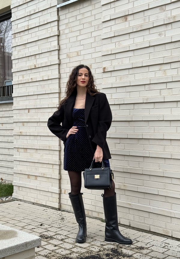 Kobieta w czarnym blezerze, granatowej sukience, czarnych rajstopach i wysokich kozaczkach stoi, trzymając czarną torebkę na tle białej ceglanej ściany.