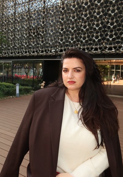 Mujer con cabello largo y oscuro, vestida con una blusa blanca y un blazer marrón, de pie en una terraza de madera frente a un edificio moderno con una fachada de patrón circular.