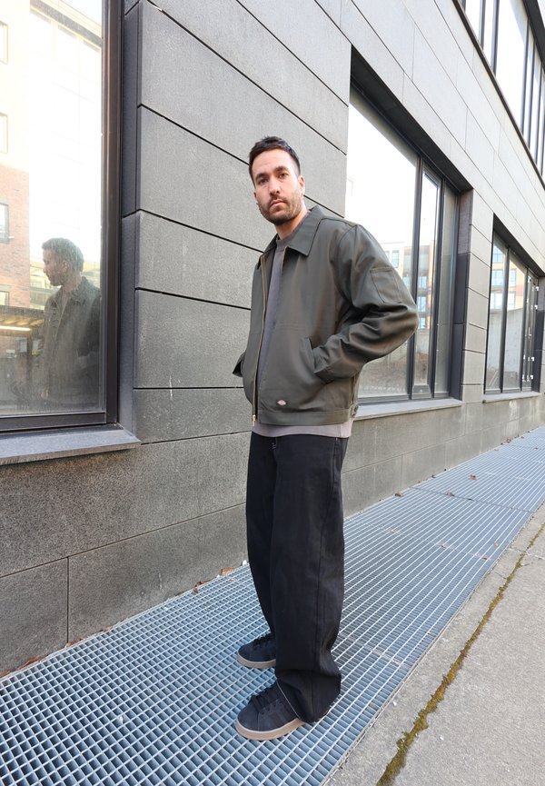 Un homme portant une veste verte et un pantalon noir se tient sur une grille métallique à côté d'un bâtiment gris avec de grandes fenêtres, les mains dans les poches, regardant devant lui.