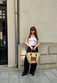 Γυναίκα με γυαλιά ηλίου, φορώντας ένα άσπρο τοπ και μαύρο παντελόνι, κρατά μια μεγάλη τσάντα από ψάθα και στέκεται μπροστά σε έναν μπεζ αστικό τοίχο.