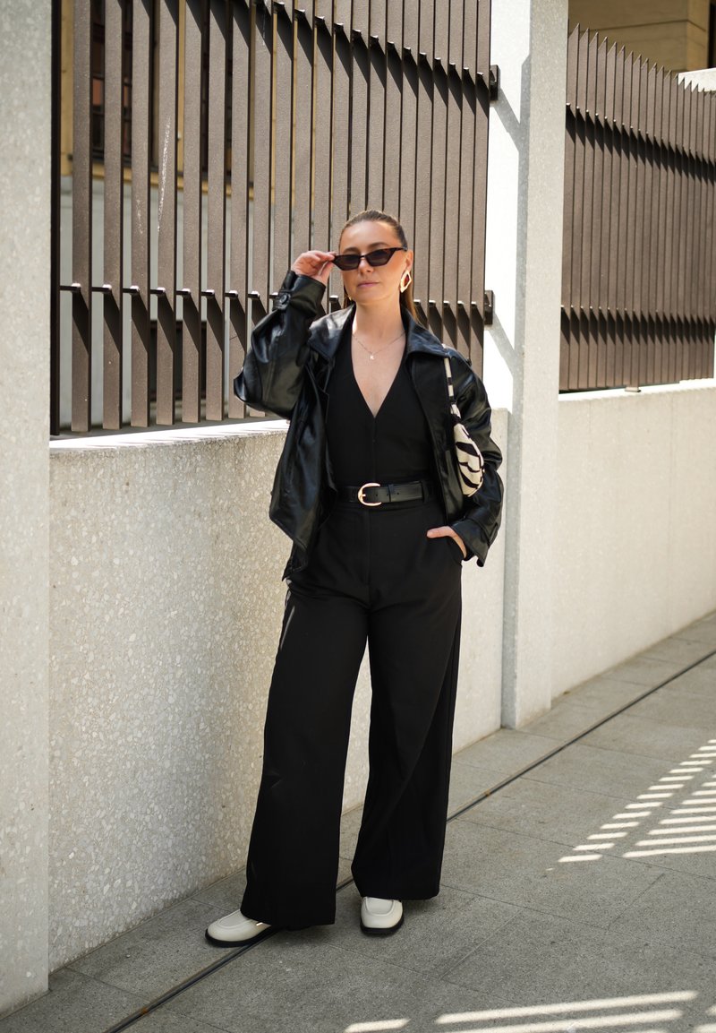 Femme portant une veste en cuir noire, un pantalon noir à jambes larges, des chaussures blanches, des lunettes de soleil et tenant un sac à motif noir et blanc près d’un mur en béton.