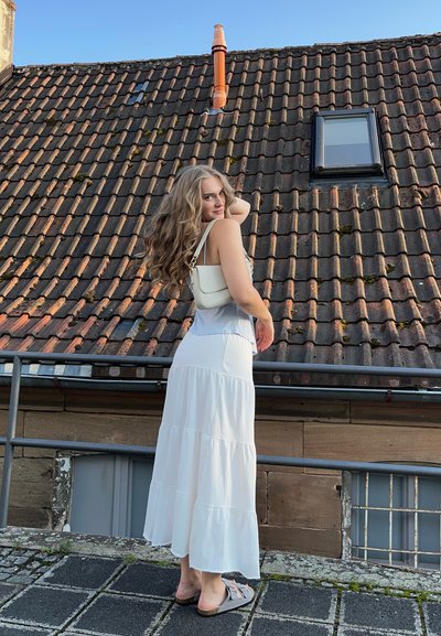 Falda maxi blanca con volantes, combinada con una blusa azul claro y sandalias grises. El fondo presenta un tejado de tejas y una pequeña ventana.