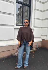 Donna che indossa un maxi maglione marrone scuro, jeans blu, scarpe marroni, occhiali da sole, tiene una piccola borsa marrone, in piedi accanto a un edificio bianco.