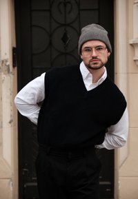 Črn pleteni brezrokavnik preko bele srajce s ovratnikom; siva kapa. Kvadratne očala, roke na bokih, stojijo pred okrašena vrata.