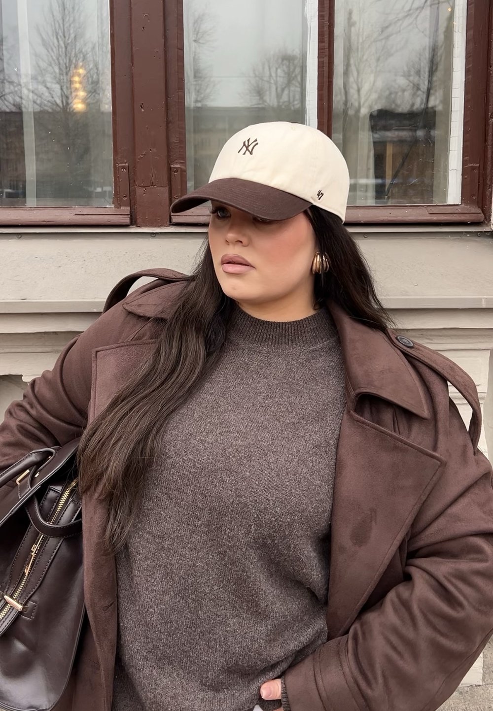 Bruine wollen trui onder een bruine trenchcoat, crèmekleurige en bruine honkbalpet, gouden oorringen en een donkere handtas.