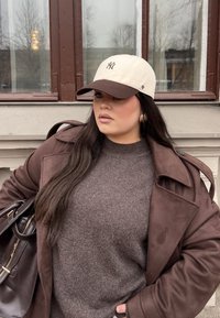 Bruine wollen trui onder een bruine trenchcoat, crèmekleurige en bruine honkbalpet, gouden oorringen en een donkere handtas.