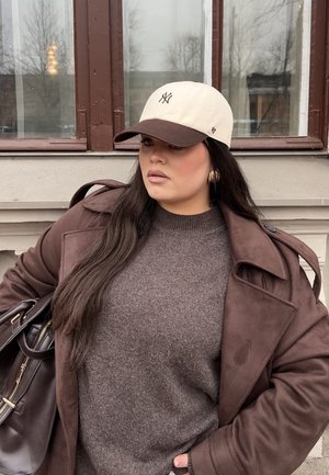 Suéter de lana marrón bajo un abrigo trench marrón, gorra de béisbol en tonos crema y marrón, pendientes de aro dorados y un bolso oscuro.