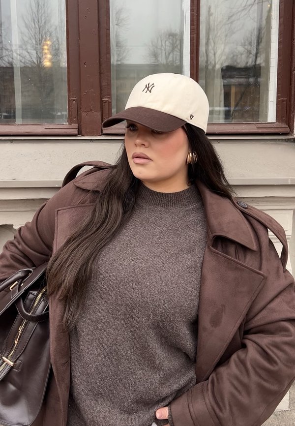 Brun ullgenser under en brun trenchcoat, kremfarget og brun baseballcap, gull øreringer i hoop-stil, og en mørk håndveske.