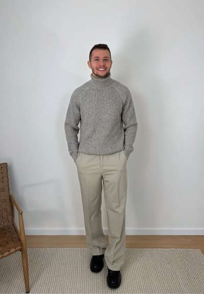 Hombre sonriente de pie con las manos en los bolsillos, vistiendo suéter gris de cuello alto, pantalones beige y zapatos negros en un entorno interior minimalista.