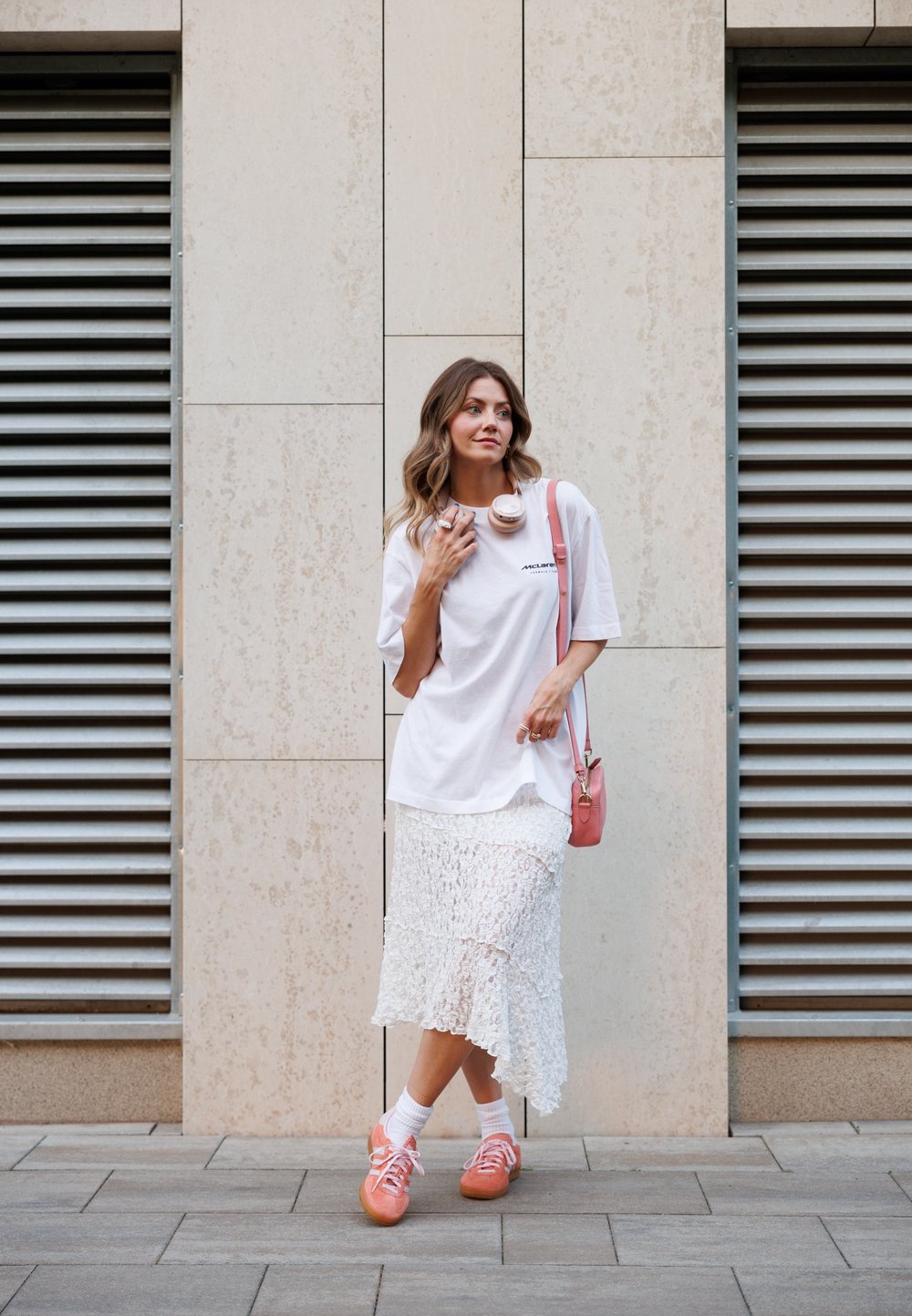 Λευκό oversized t-shirt, λευκή δαντελωτή midi φούστα, ροζ αθλητικά παπούτσια, ροζ τσάντα χιαστί και ροζ ακουστικά. Υφές και επίπεδες σχεδίες.