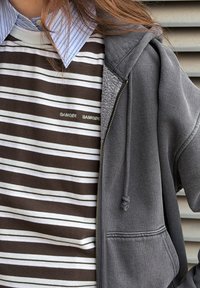 Sudadera con cremallera gris sobre una camiseta de manga larga a rayas marrón y blanca. La camiseta presenta un logotipo, y la sudadera tiene un bolsillo de tela texturizada.