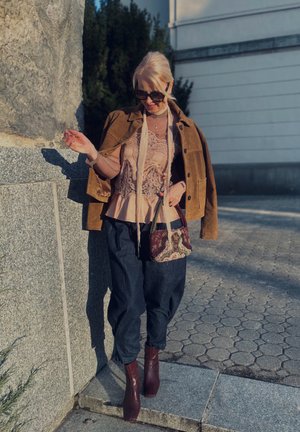 Mujer con chaqueta de ante marrón, blusa de encaje, pantalones oscuros, gafas de sol y botas granate está de pie al aire libre junto a una pared de piedra bajo la luz del sol.