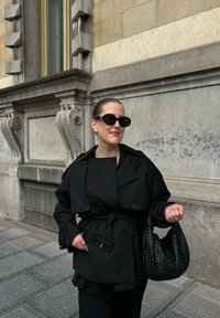 Kobieta w czarnym płaszczu trench oraz okularach przeciwsłonecznych, trzymająca dużą, plecioną czarną torebkę, stojąca na brukowanej ulicy obok kamiennego budynku.