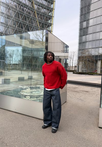 Hombre con suéter rojo y pantalones vaqueros anchos de pie al aire libre cerca de una estructura de vidrio, con edificios modernos y árboles sin hojas en el fondo.