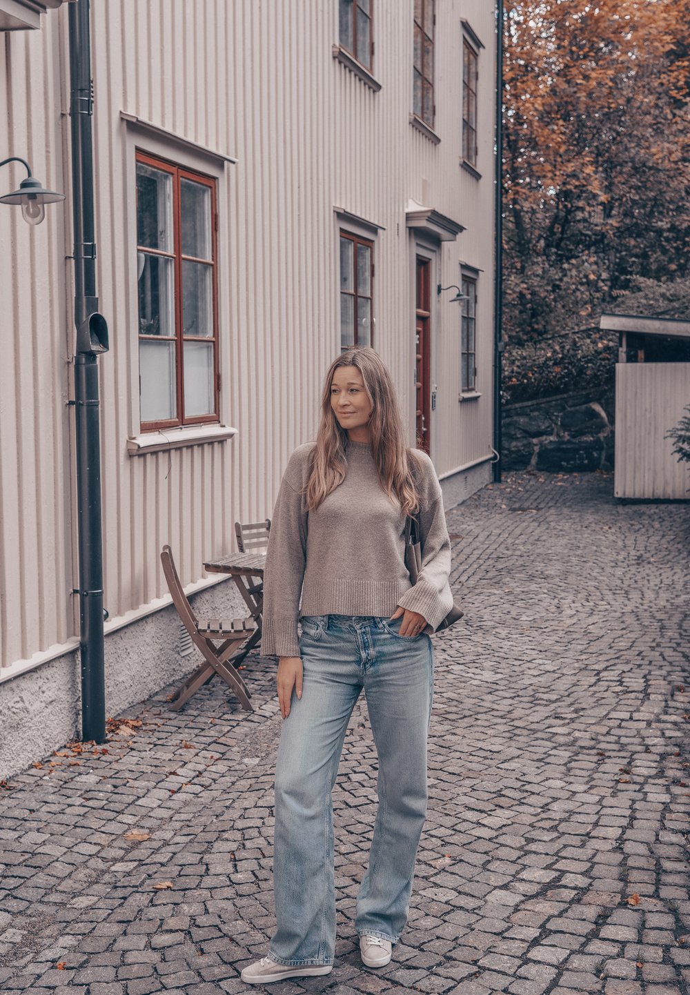 Jovem mulher com suéter cinza e jeans, em pé numa rua de calçada, ao lado de um edifício branco de madeira, com árvores de outono ao fundo.